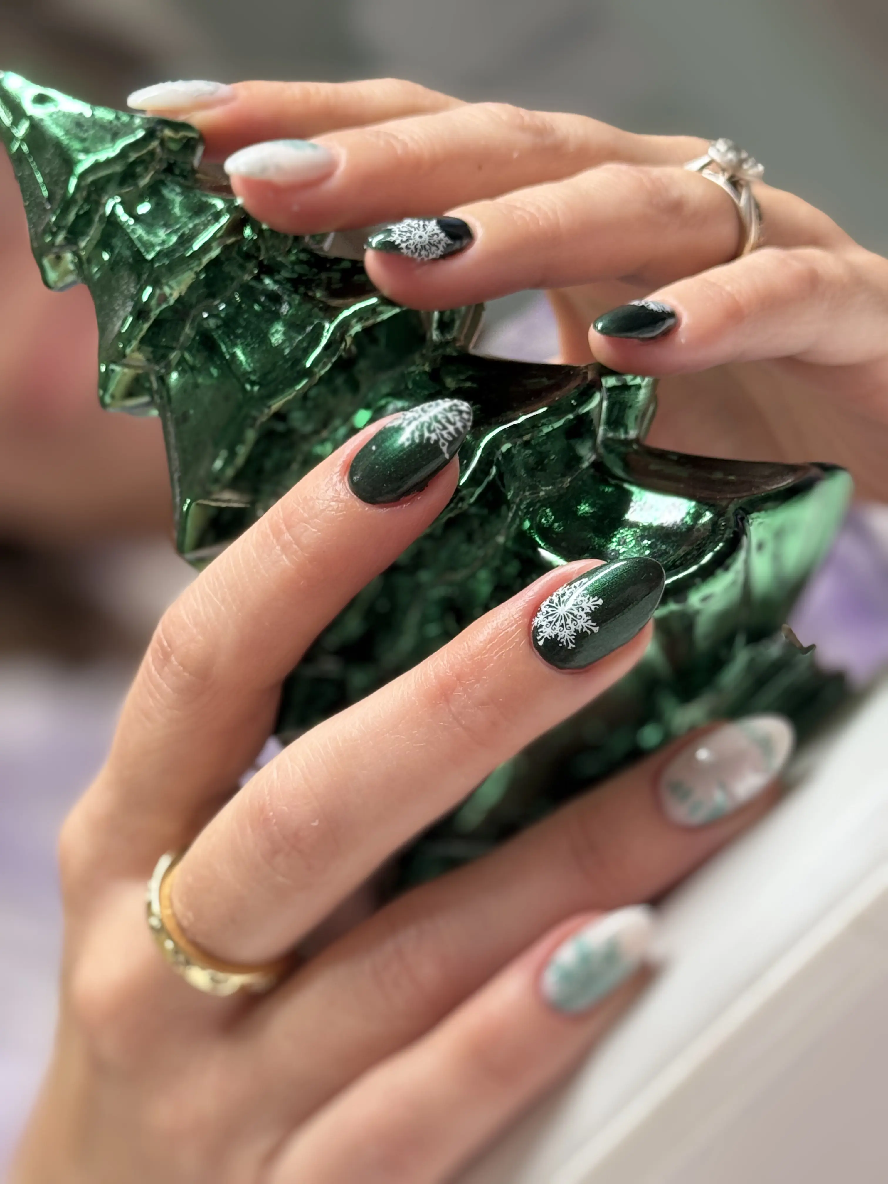 Almond nails in dark green gel polish with hand-painted white snowflakes, holding a green Christmas tree ornament