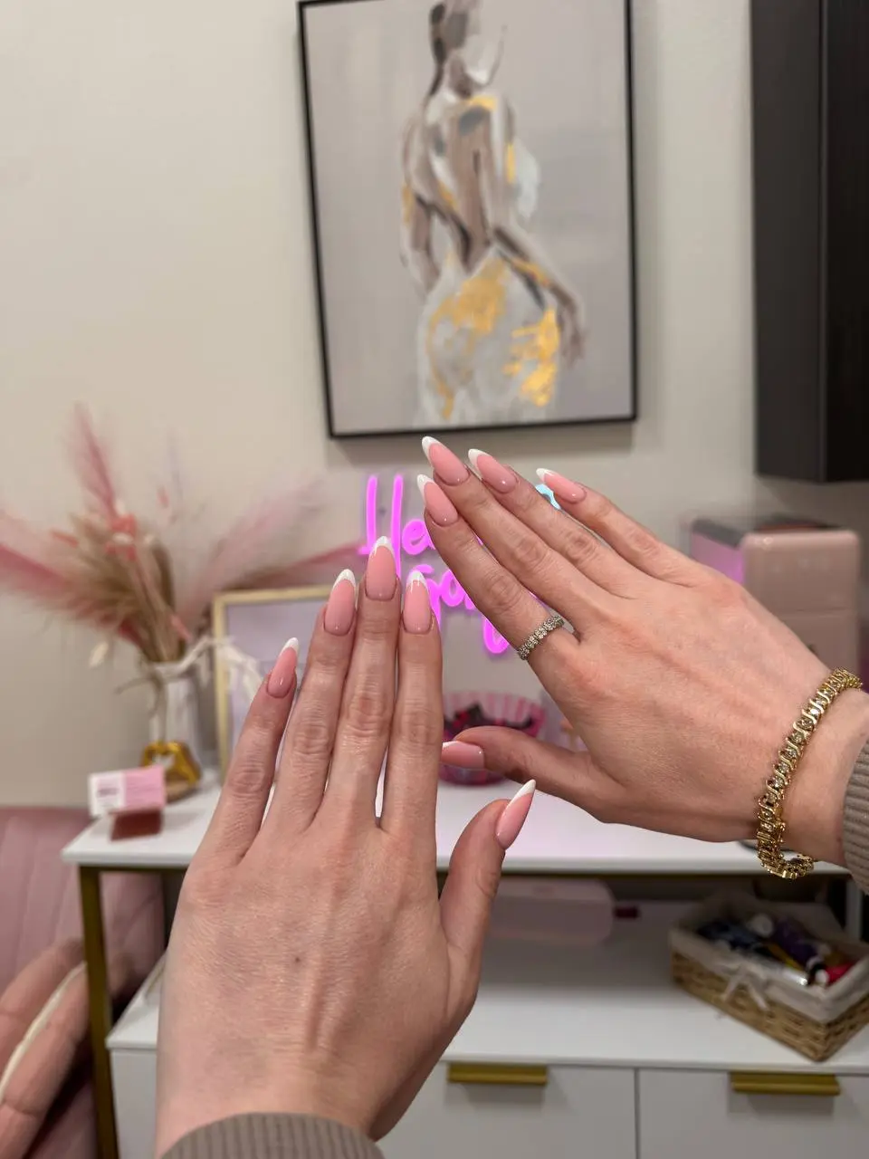 Two hands with long almond French manicures and white tips, in front of the studio's pink 'Hello Gorgeous' neon sign